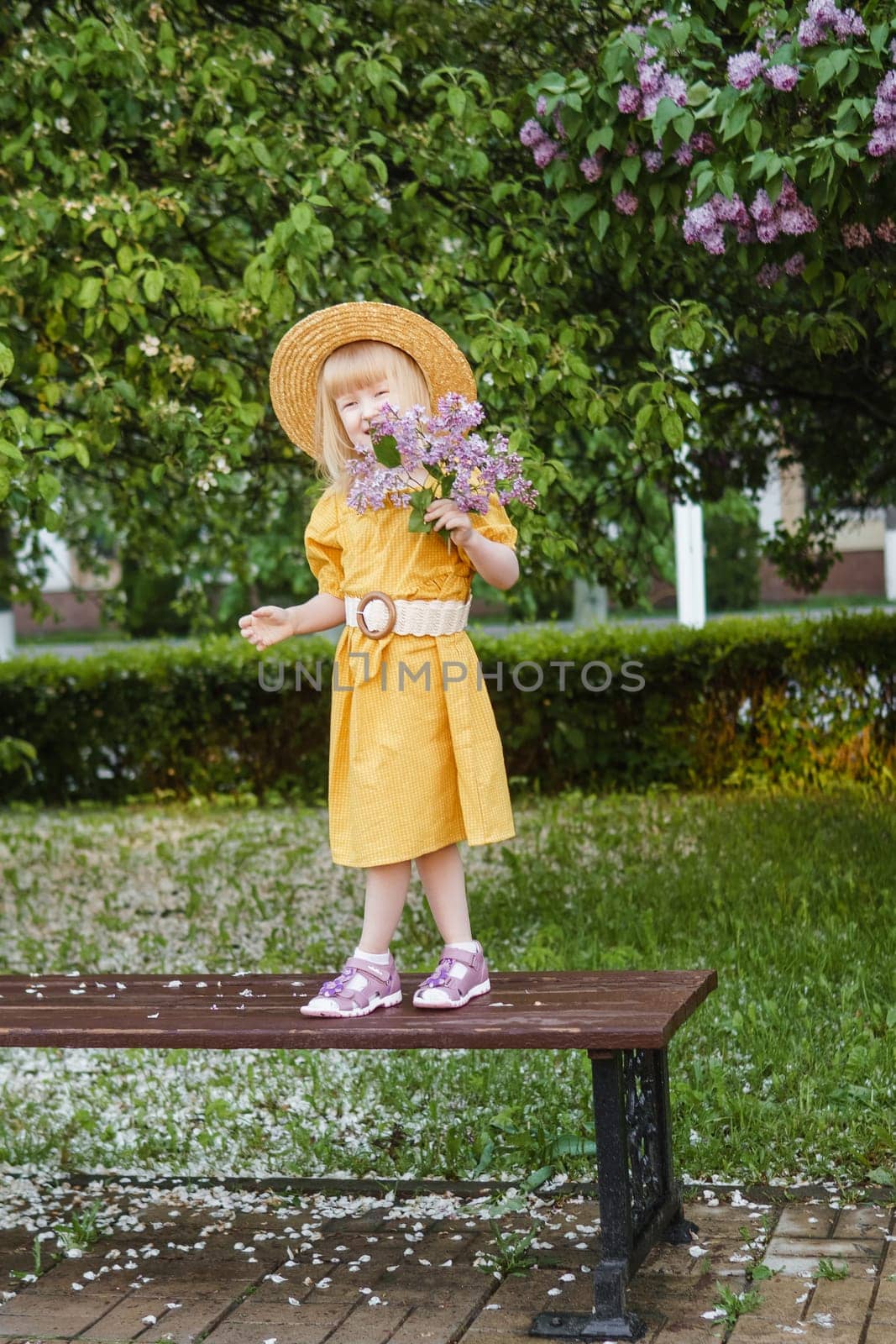 A little girl in a yellow dress and straw hat wearing a bouquet of lilacs. A walk in a spring park, blossoming lilacs