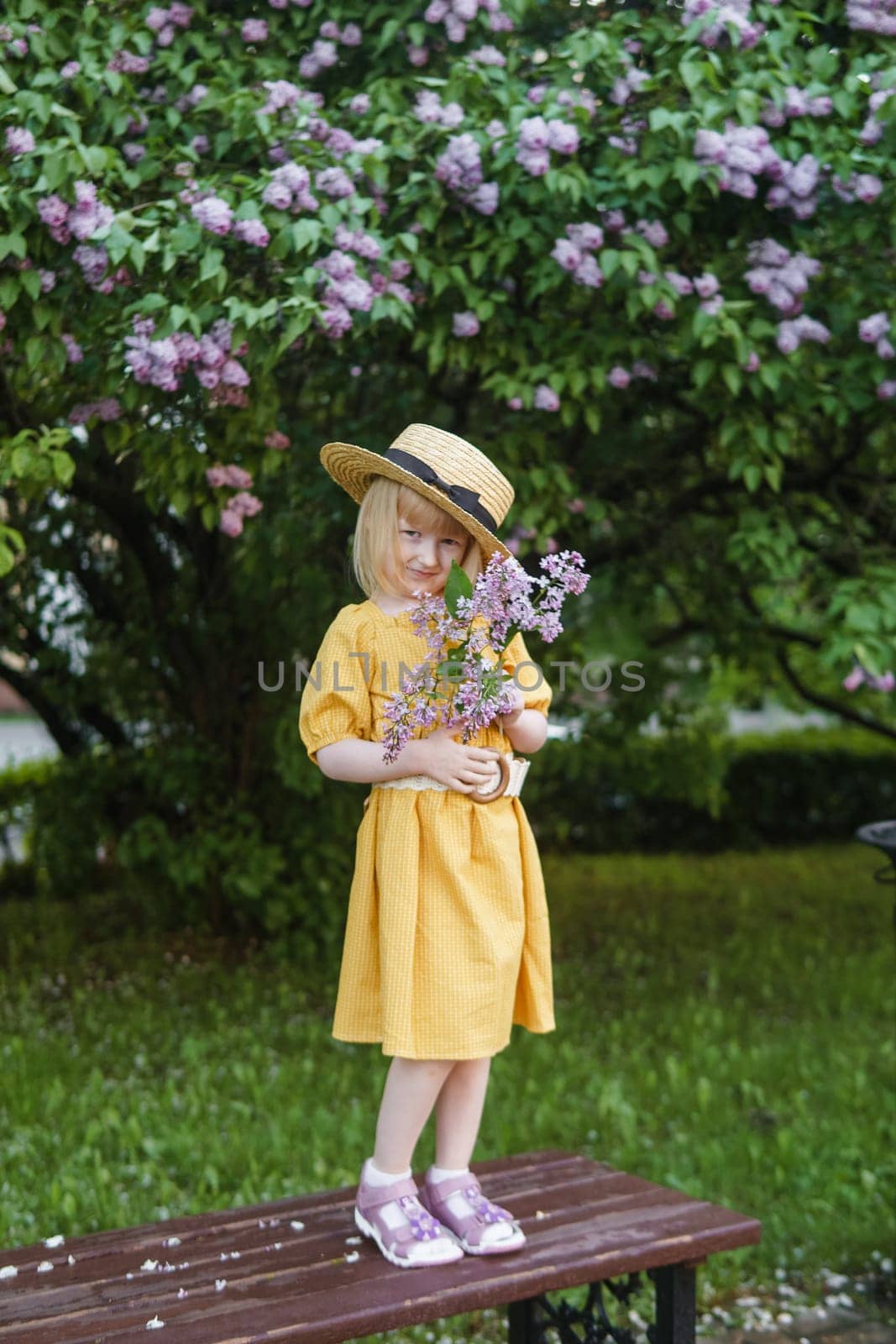 A little girl in a yellow dress and straw hat wearing a bouquet of lilacs. A walk in a spring park, blossoming lilacs