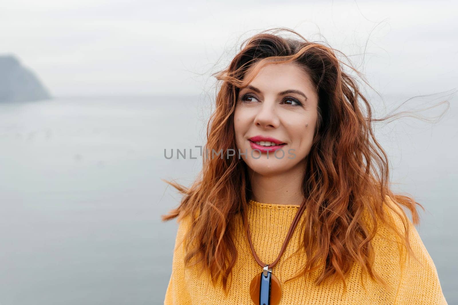 Portrait windswept hair happy woman against a backdrop of mountains and sea. Daylight illuminates the tranquil outdoor setting.