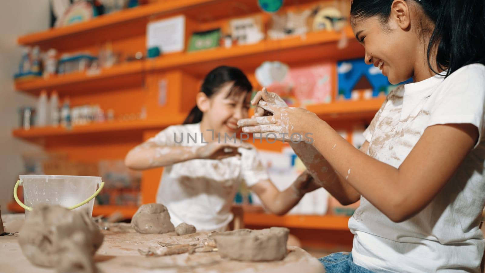 Happy multicultural girl and friend put the clay on the each other muddy shirt at art lesson. Diverse highschool student playing with clay at pottery workshop. Creative activity concept. Edification.