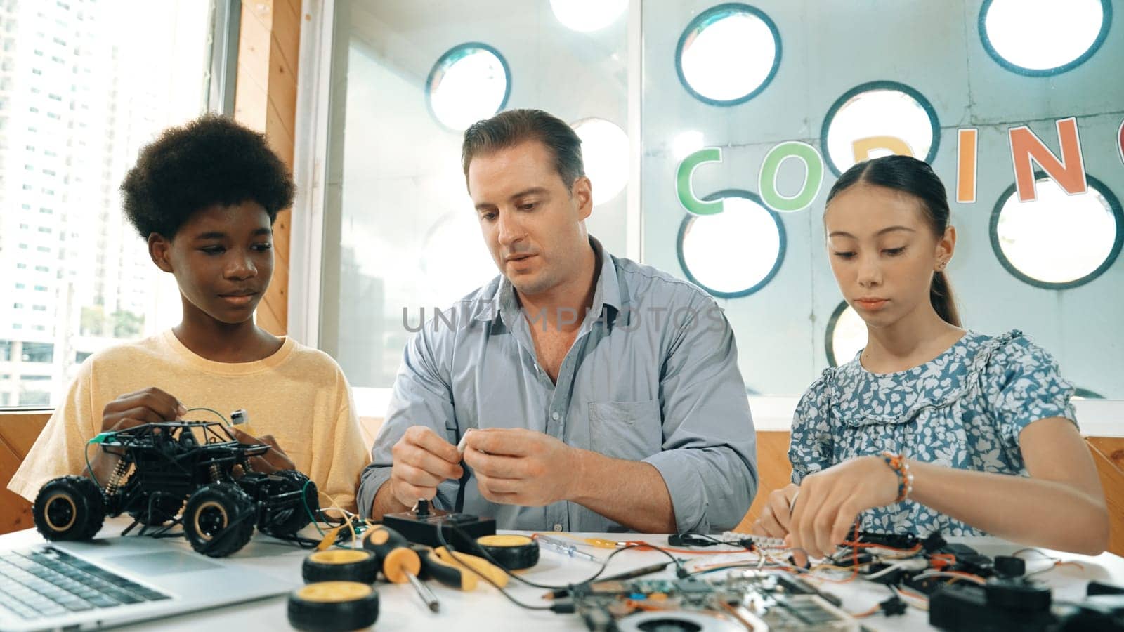 Skilled teacher teaching diverse students fixing car model by using wire and electronic equipment. Girl studying about using electrical tool to check mother board while looking at laptop. Edification