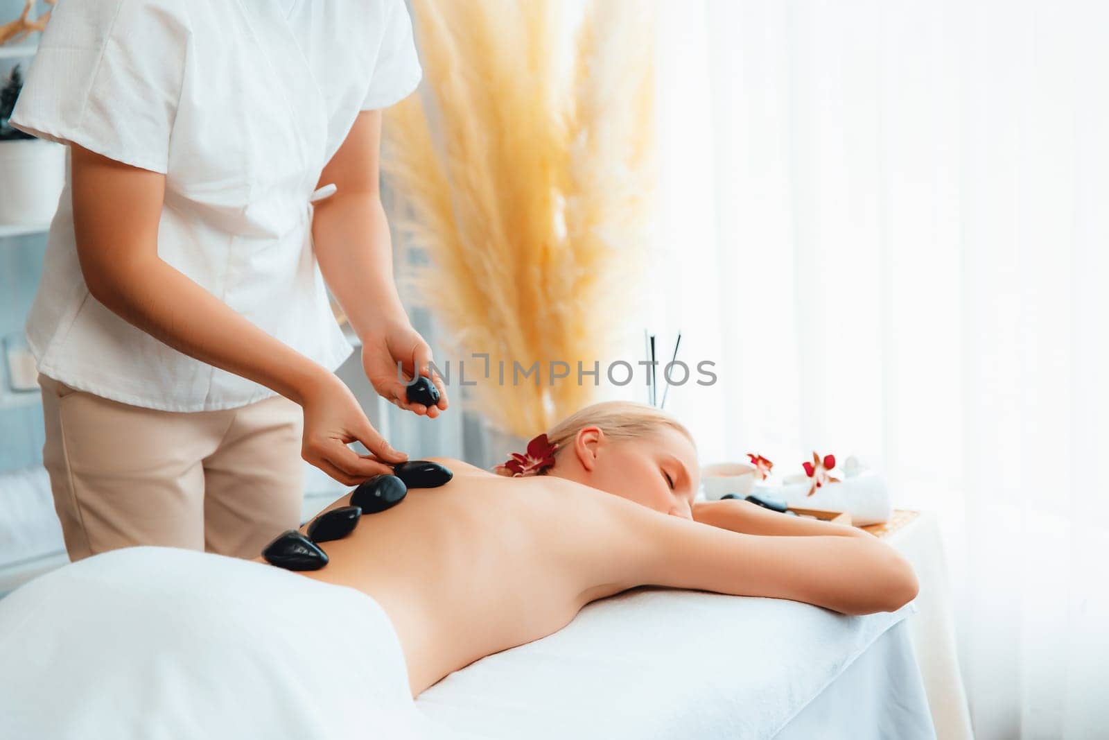 Hot stone massage at spa salon in luxury resort with day light serenity ambient, blissful woman customer enjoying spa basalt stone massage glide over body with soothing warmth. Quiescent