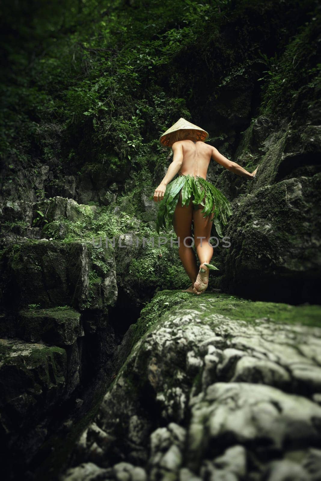 A naked sexy girl in a triangular Asian hat and a homemade cape made of leaves walks along the canyon of mountain river