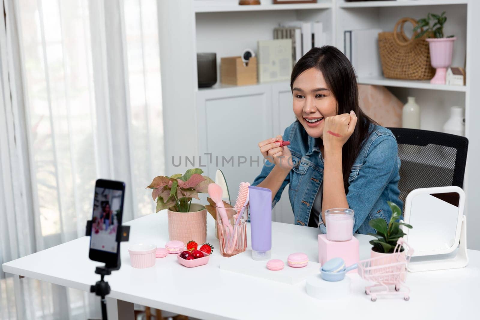 Young beautiful Asian showing cosmetic makeup lipstick matte pastel color testing in bestseller collection on arm test promotion on social media online recording smartphone at modern room. Stratagem.