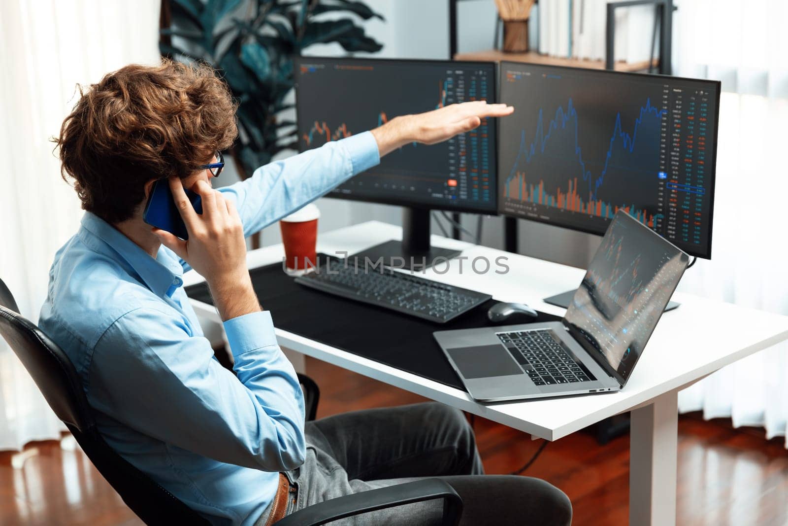 Successful young investor stock trader calling on smartphone to business partner with dynamic financial exchange investment laptop screen in current market graph in real time at modern office. Gusher.
