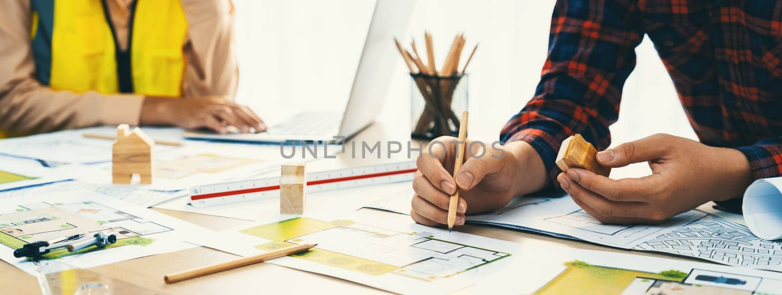 Professional architect and engineer collaborate to design eco-friendly house on meeting table with blueprint, wooden house block and tree model scatter around. Closeup. Delineation.