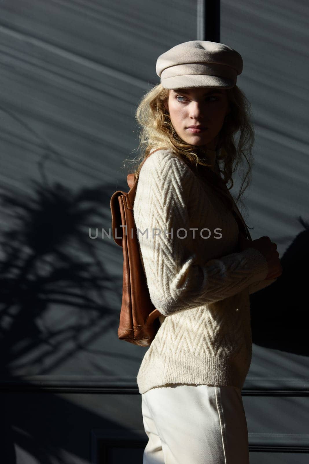 Studio portrait of beautiful woman with a curly blond hair holding brown backpack, posing on gray background. by Ashtray25