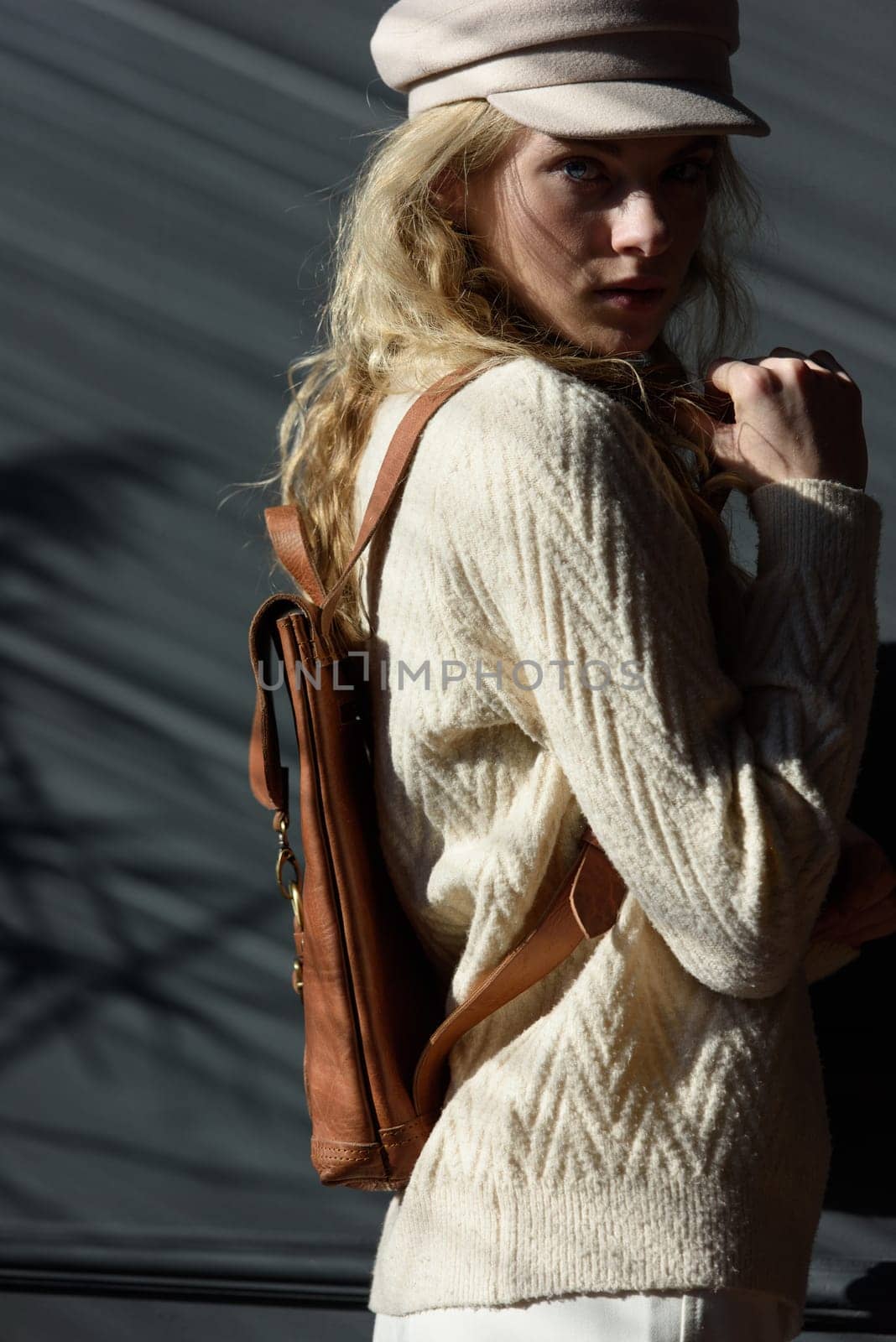 Studio portrait of beautiful woman with a curly blond hair holding brown backpack, posing on gray background. by Ashtray25