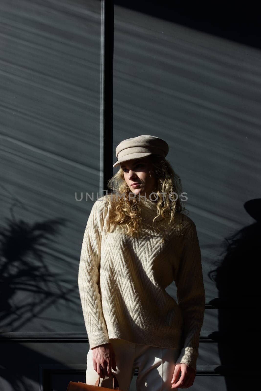 Studio portrait of beautiful woman with a curly blond hair, posing on gray background. Model wearing stylish cap, sweater and classic trousers