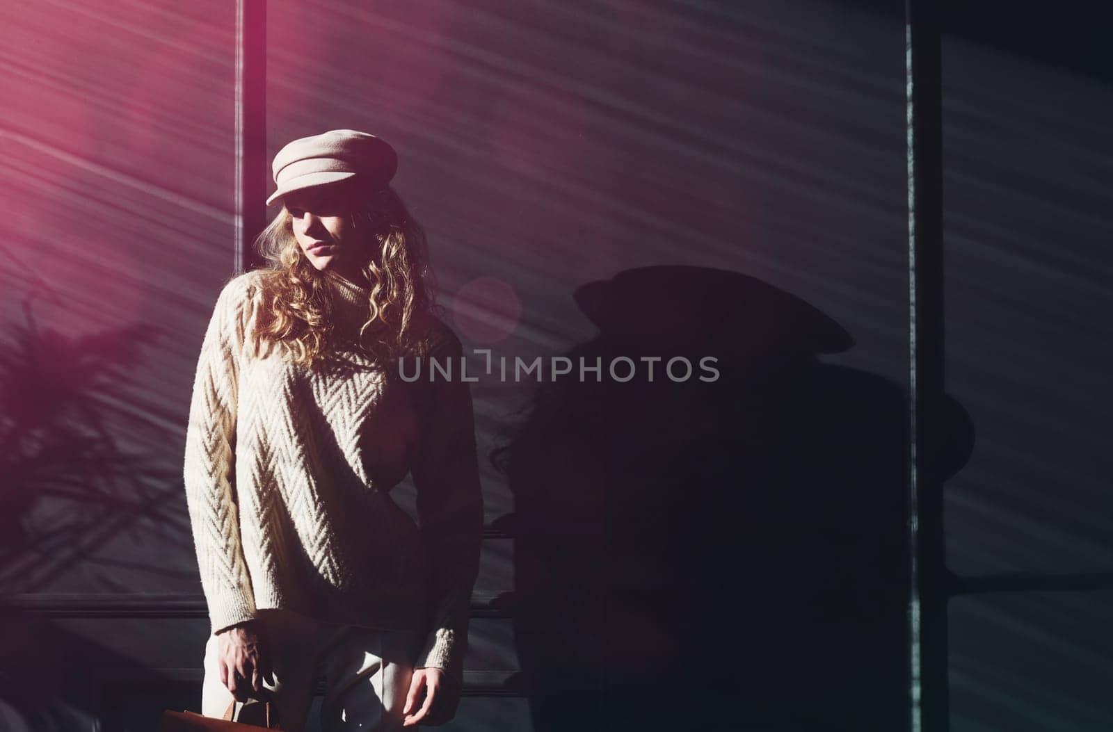 Studio portrait of beautiful woman with a curly blond hair, posing on gray background. Model wearing stylish cap, sweater and classic trousers