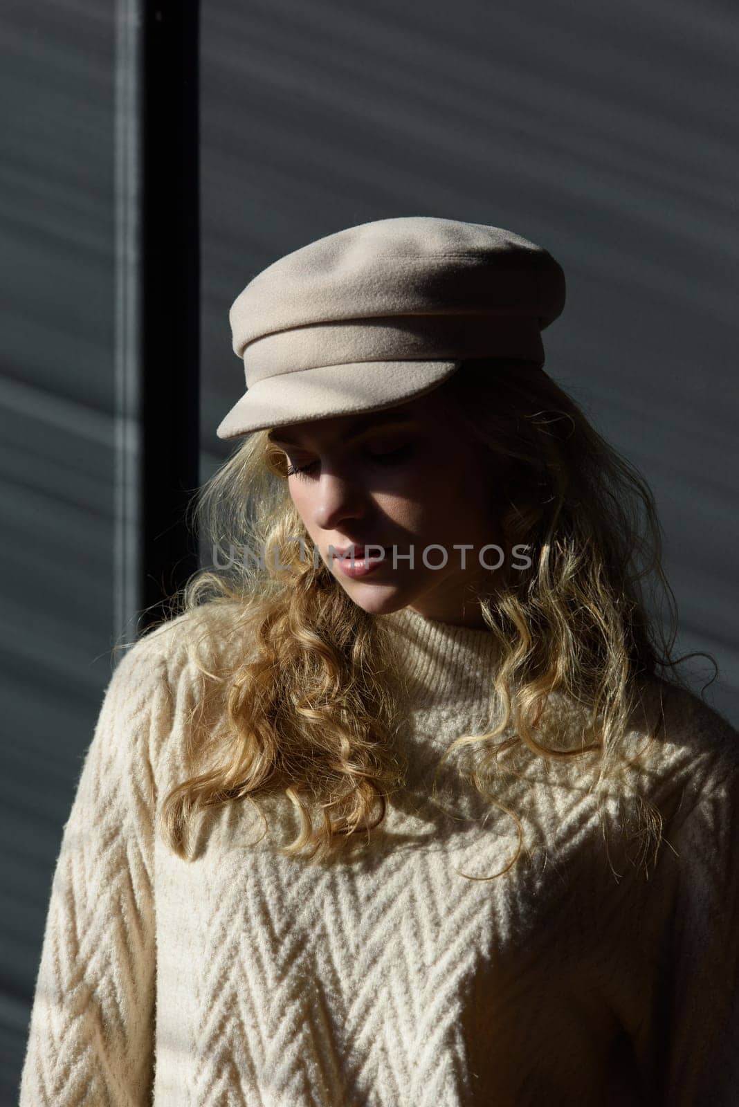 Studio portrait of beautiful woman with a curly blond hair, posing on gray background. Model wearing stylish cap, sweater and classic trousers