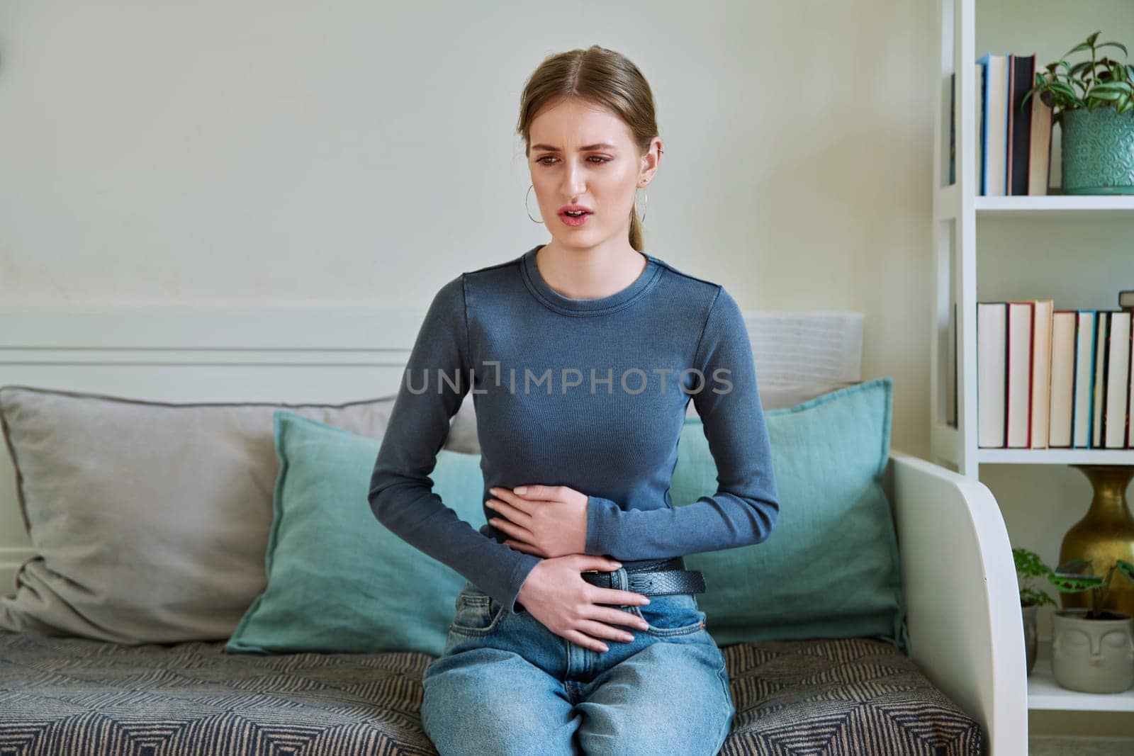 Young teenage female experiencing abdominal pain, sitting on couch at home by VH-studio