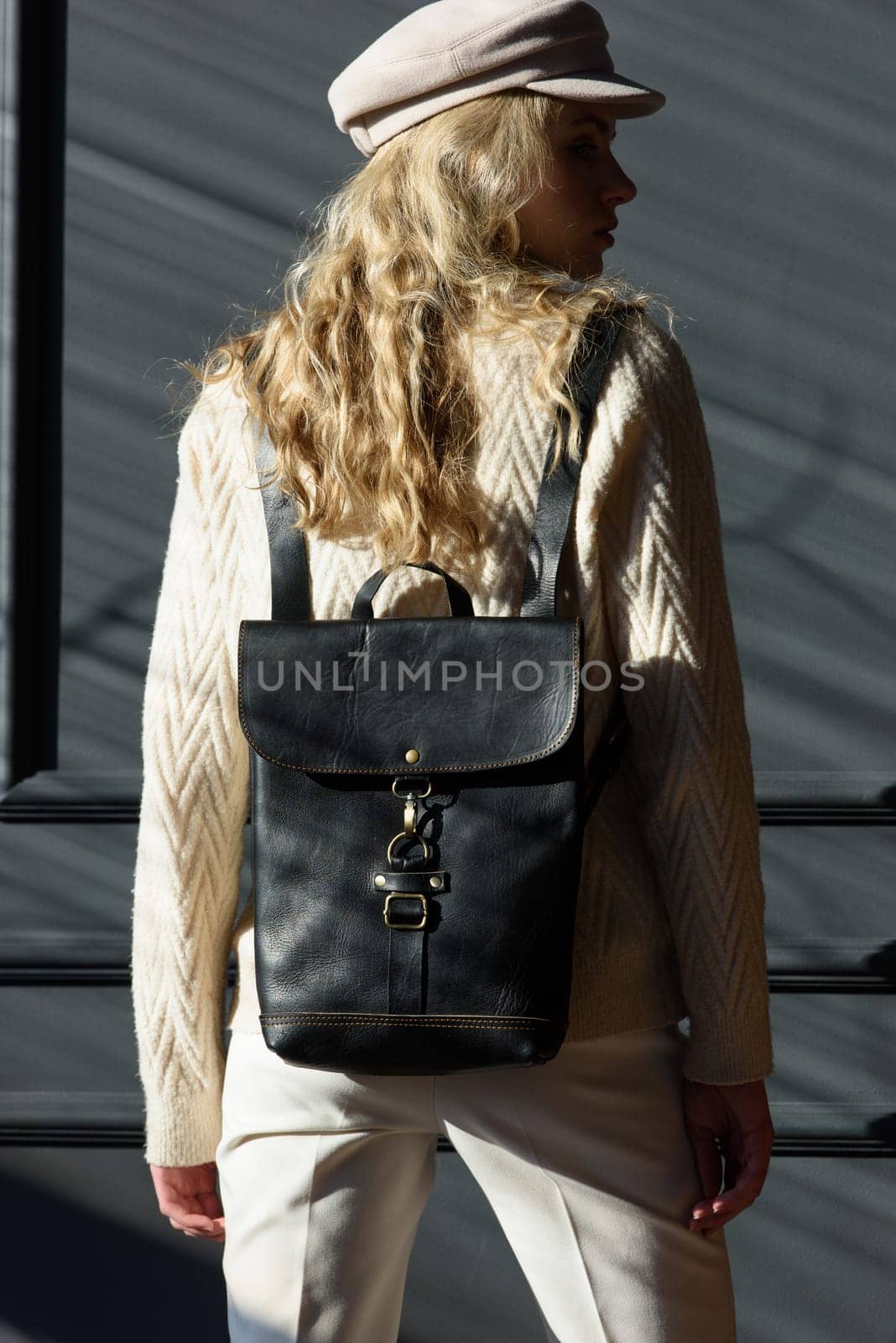 Studio portrait of beautiful woman with a curly blond hair holding brown bag, posing on gray background. Model wearing stylish cap, sweater and classic trousers