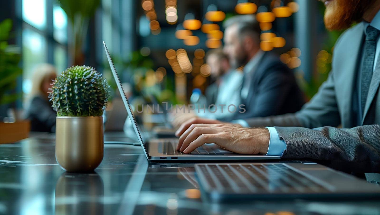 A man in a suit is typing on his laptop computer at a table. There is a houseplant and flowerpot on the table. The room has elegant flooring and tableware set up for an event