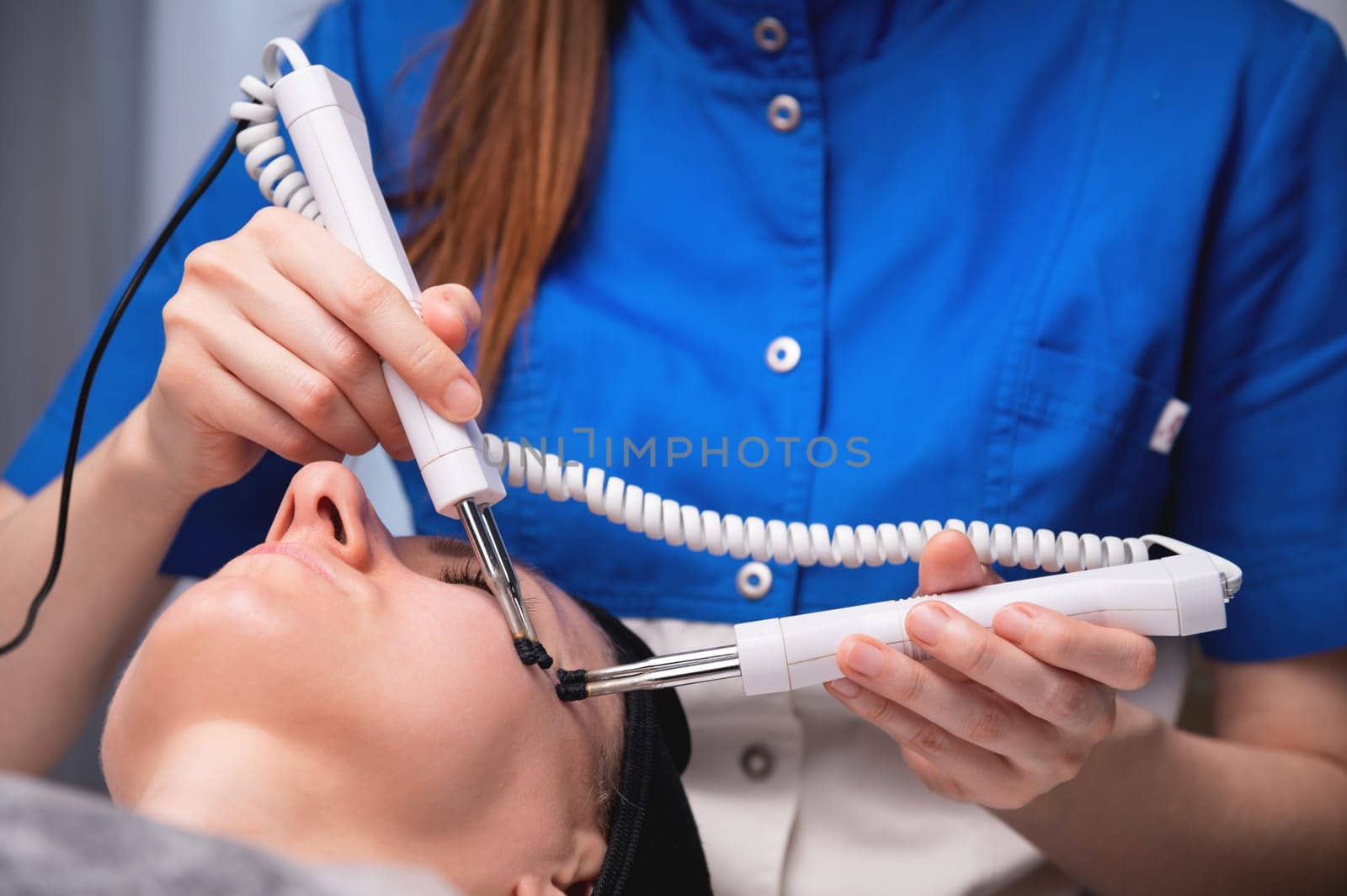 A middle-aged woman undergoes electrotherapeutic facial rejuvenation procedures in a cosmetologist's office. by yanik88