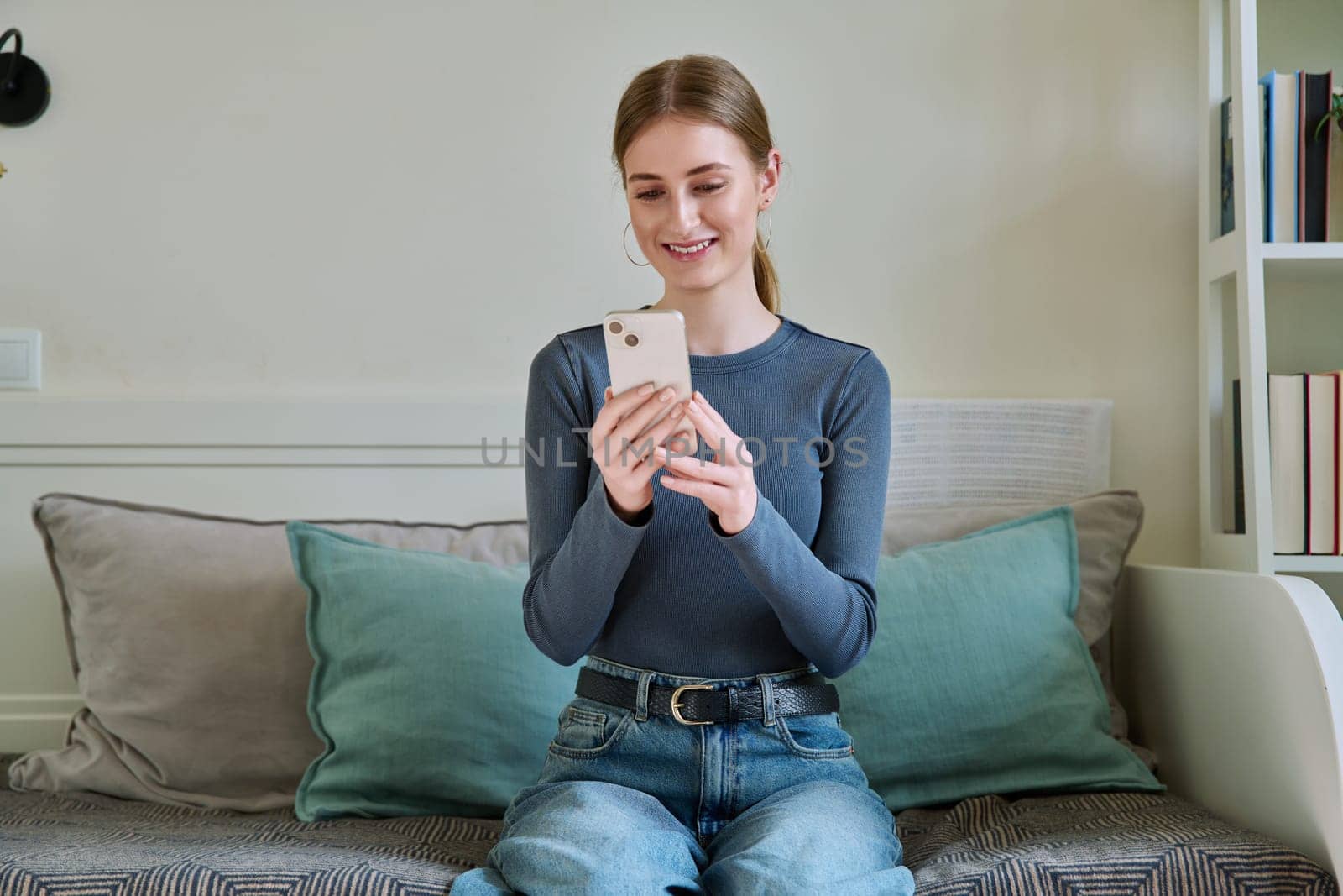 Happy female teenager using smartphone, sitting on couch at home by VH-studio