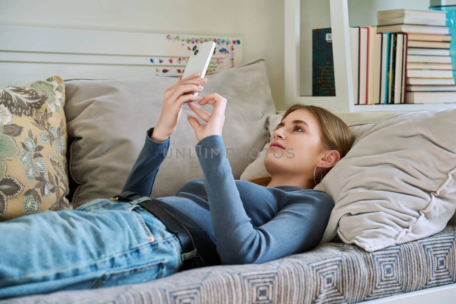 Happy smiling relaxed young female teenager lying on couch at home using smartphone for leisure communication. Youth, lifestyle, digital online technology, gadgets, internet