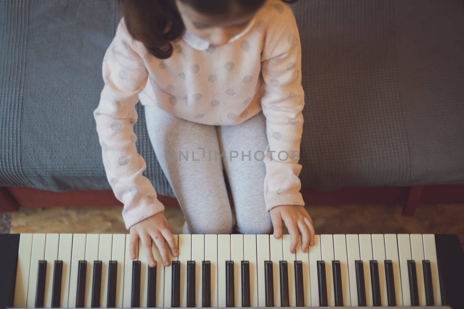 A beautiful little caucasian brunette girl with two ponytails enthusiastically presses the keys on an electric piano while sitting on a sofa in the room, close-up top view. Music education concept.