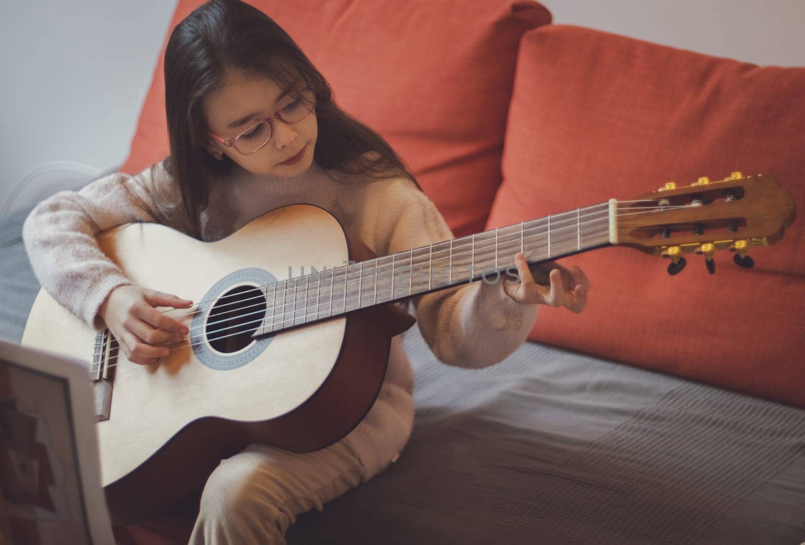 A beautiful little caucasian brunette woman with flowing long hair in glasses holds a guitar and plucks the strings with her fingers, studying notes while sitting on a sofa in the room, top view close-up. Music education concept.