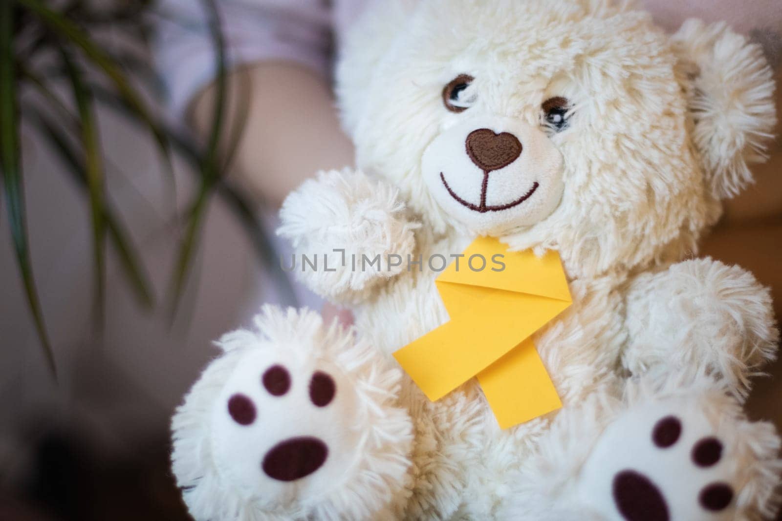 Little caucasian brunette female is holding in her hands a teddy bear with a yellow ribbon tie on a white wall background with a flower,close-up side view with depth of field. World childhood cancer day concept.