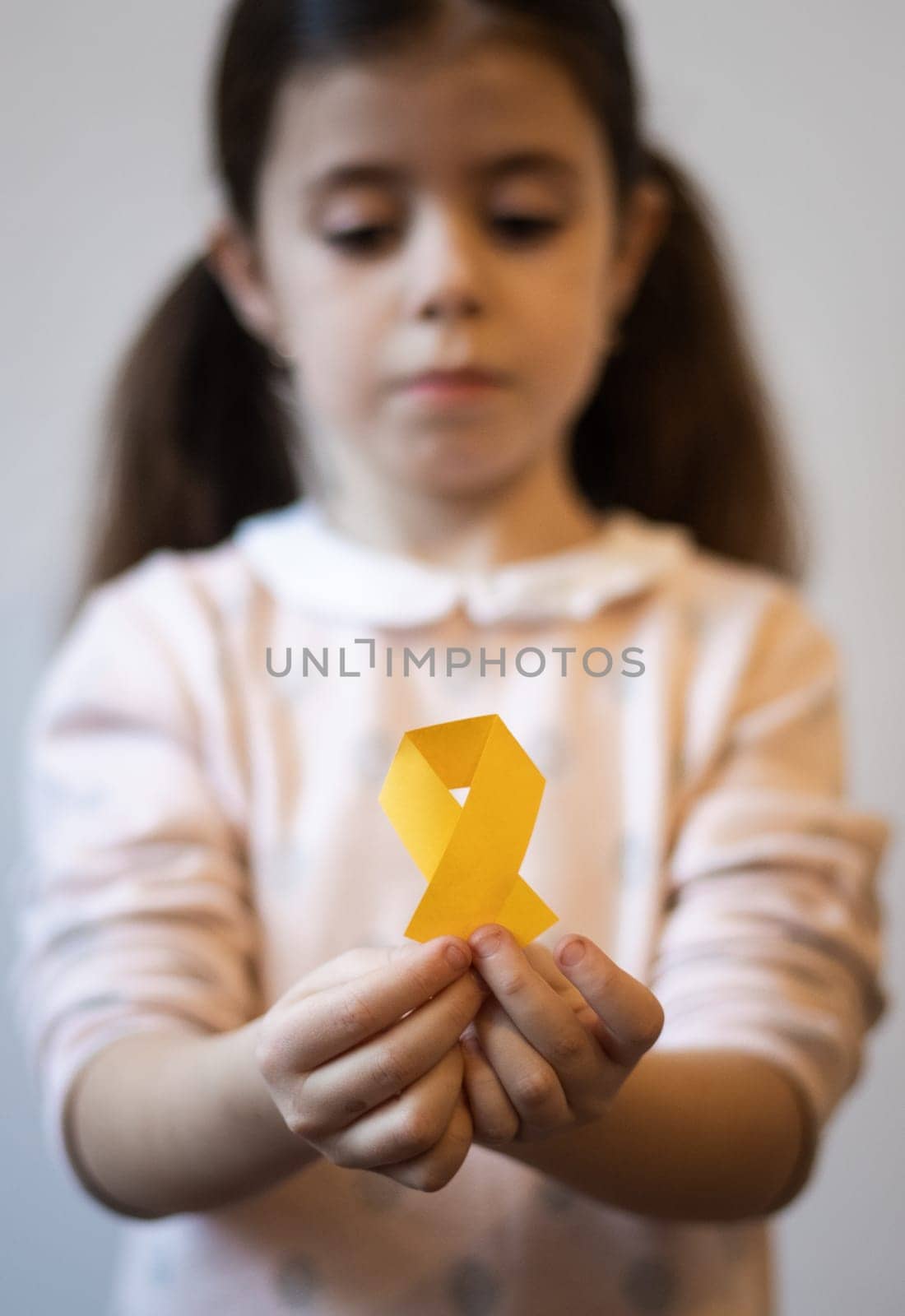 Beautiful little caucasian brunette girl with a sad face is holding out a yellow paper tape in her hands on a white background,close-up side view with depth of field. World childhood cancer day concept.