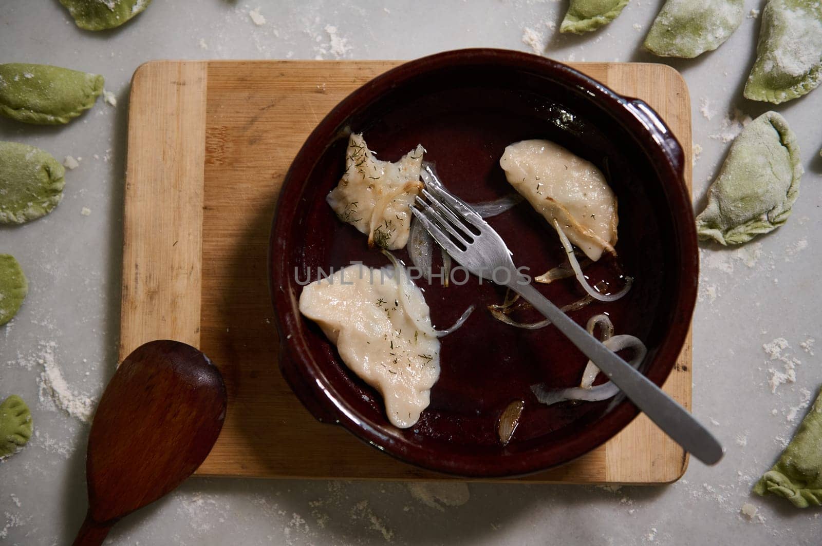 Dirty crockery and clay plate with the rest of unfinished lunch of varennyki with roasted onion on kitchen table with raw molded dumplings. Food blogging. Still life. View from above Leftover of lunch