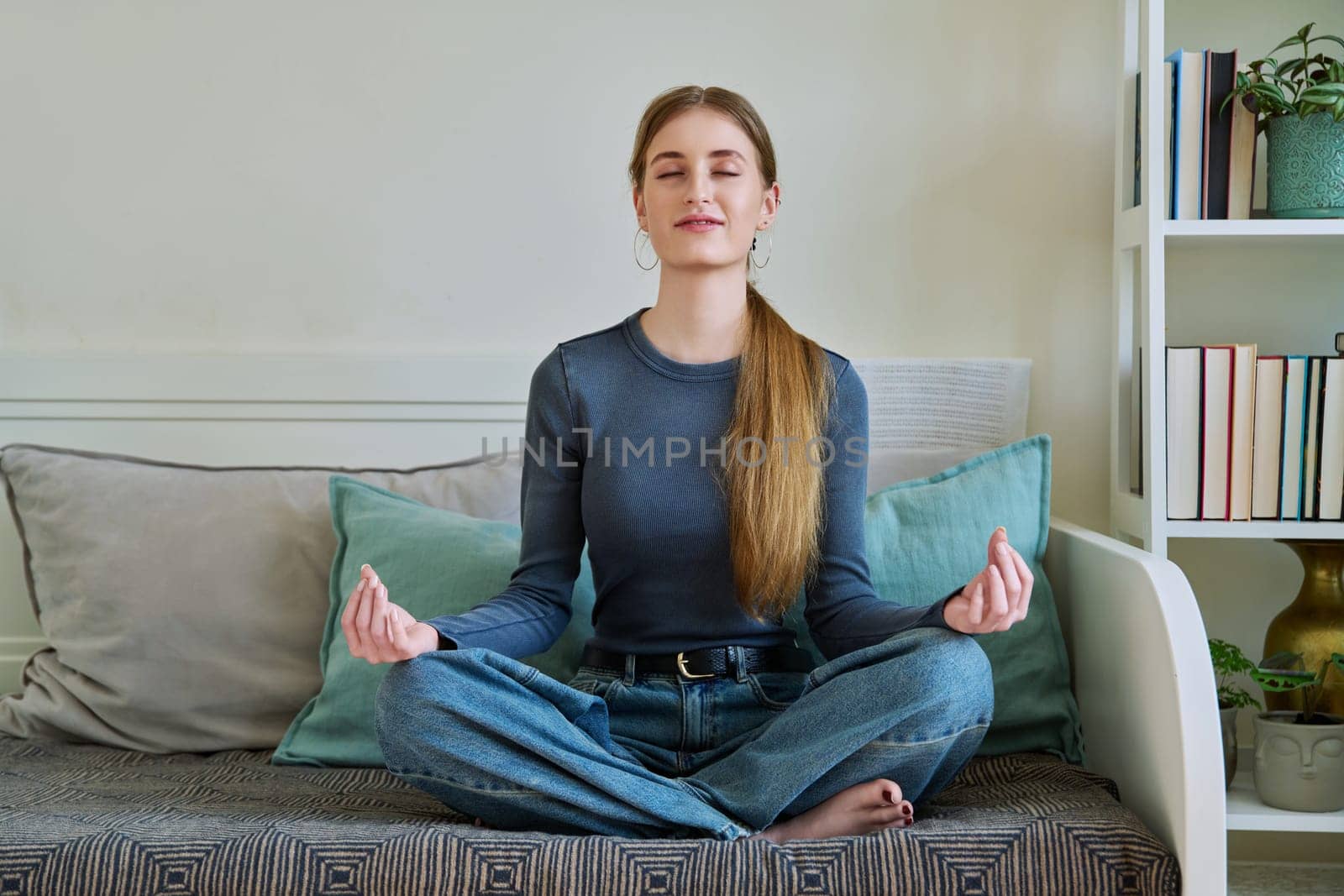 Young teenage relaxed female sitting in lotus position, meditating at home by VH-studio