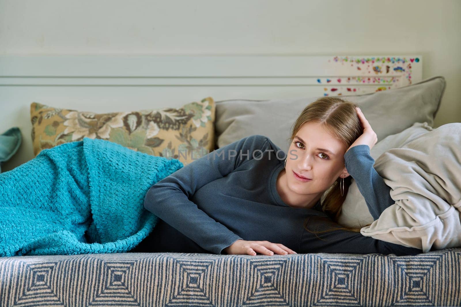 Beautiful smiling calm young teenage female relaxing lying under blanket at home on couch, looking at camera.