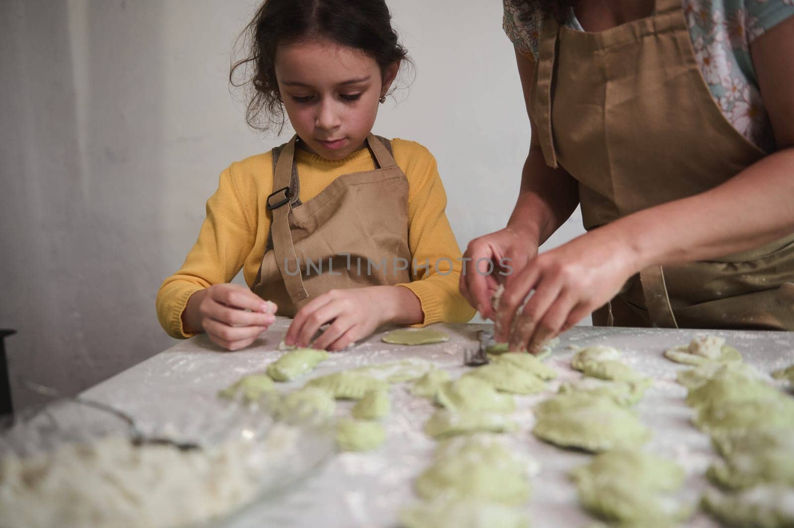Lovely little girl in beige chef apron, standing at floured kitchen table, helping her mother to stuff and sculpt dumplings. Mom and daughter cooking together varennyky according to traditional recipe