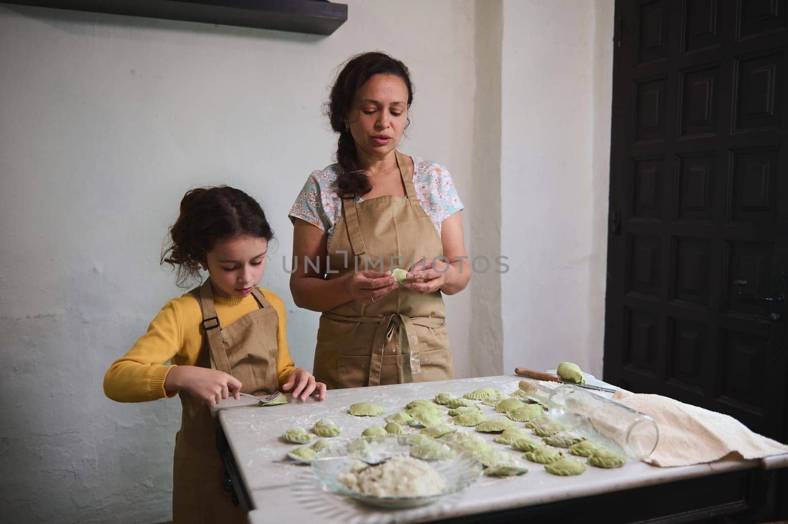 Lovely little girl in beige chef apron, standing at floured kitchen table, helping her mother to stuff and sculpt dumplings. Mom and dughter cooking together