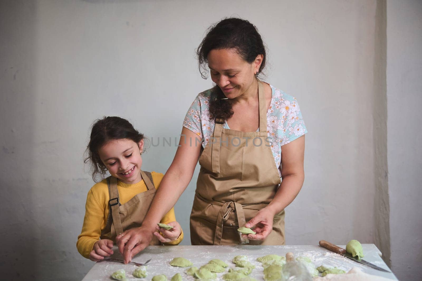 Happy mother nd daughter cooking dumplings in the rustic home kitchen of a rural house. Little daughter learning stuffing nd molding Ukrainian varennyky or Italian ravioli. People. Cuisine. Culinary