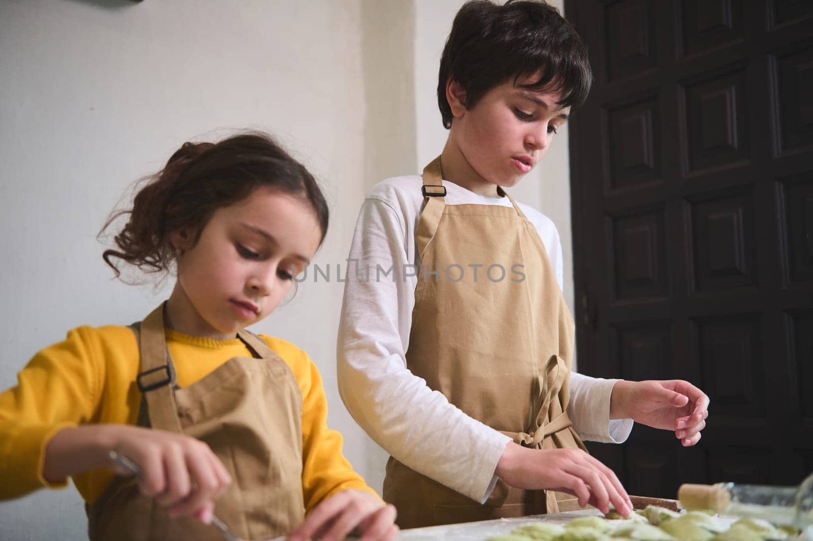 Cute kids boy and girl in the rural kitchen, sculpting dumplings from dough with mashed potatoes filling. Cooking homemade dumplings, Italian ravioli or Ukrainian varenyky according traditional recipe