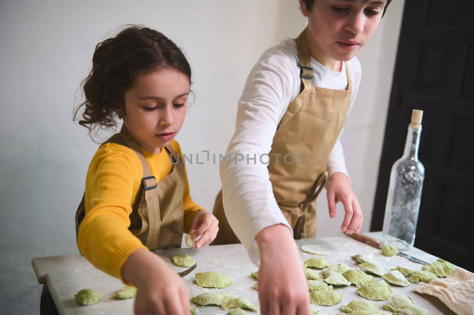 Boy and girl making dumplings, standing at floured kitchen table in the rustic kitchen interior. Cooking class for kids. Preparing ravioli, pelmeni, varennyky according to traditional recipe by artgf