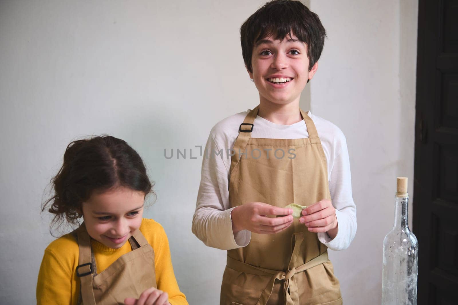 Authentic portrait of two adorable smiling kids making homemade dumplings at home kitchen, standing against white wall background by artgf
