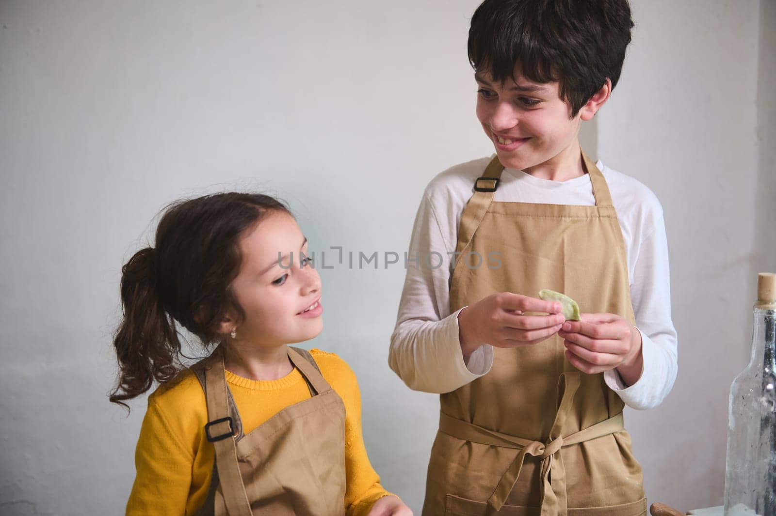 Authentic portrait of two adorable smiling kids making homemade dumplings at home kitchen, standing against white wall background