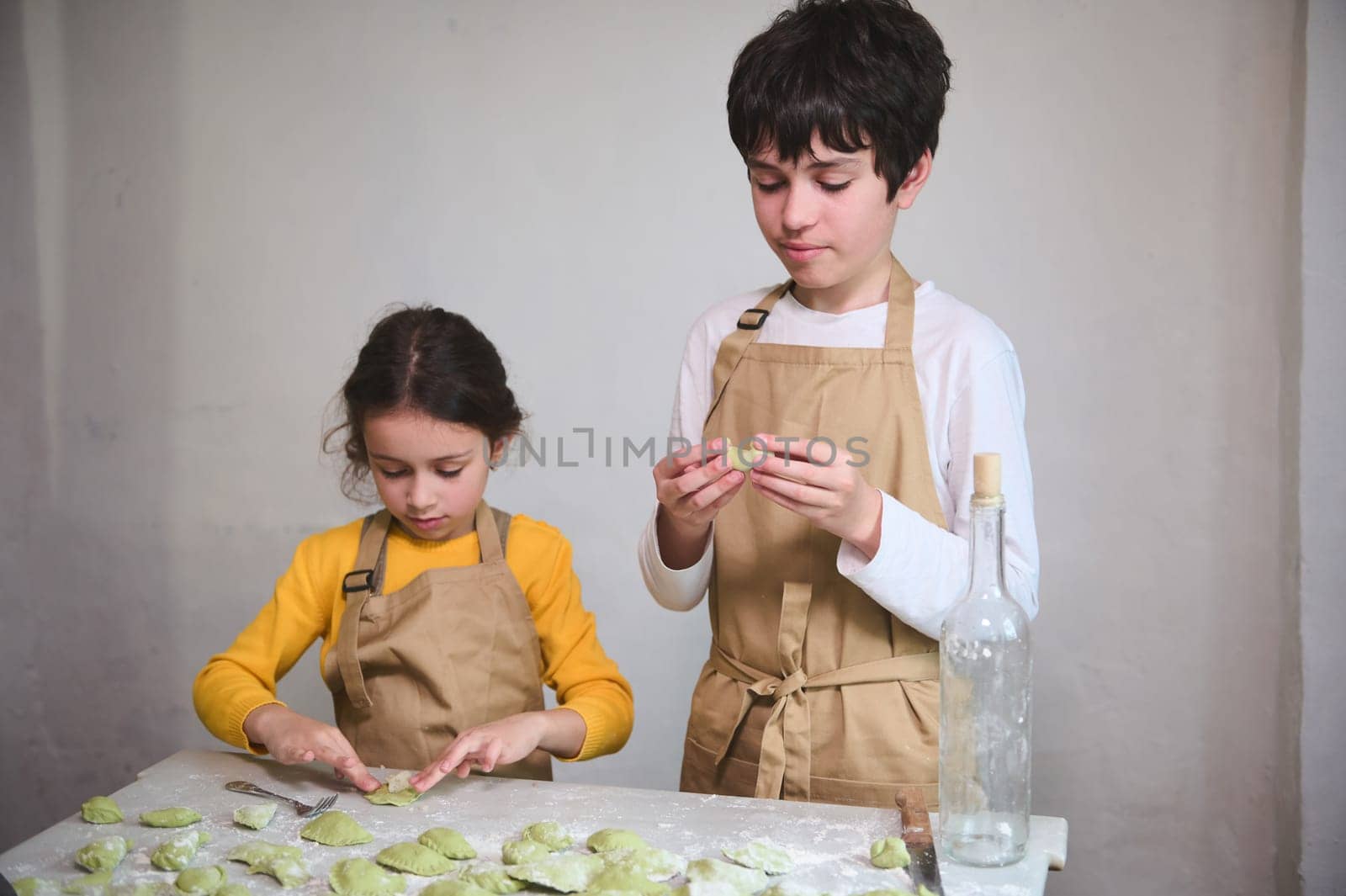 Cooking class for kids. Two diverse kids, a boy and a girl preparing family dinner, standing at floured kitchen table and modeling dumplings or Ukrainian varennyky in the rural house kitchen interior