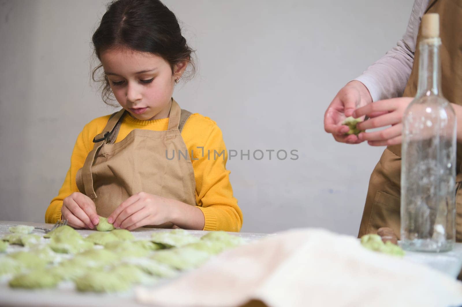 Children learn making dumplings during a cooking class. Preparing homemade varennyky according to traditional Ukrainian recipe. Close-up little kid girl sculpting dumpling, standing at floured table