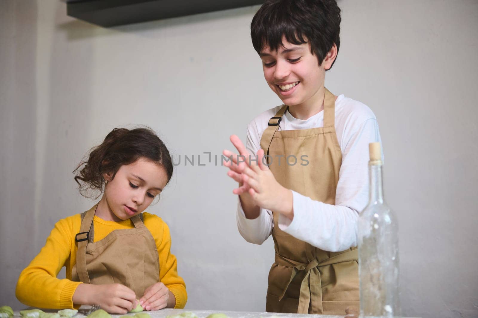 Cute children in beige chef apron, preparing dumplings during a cooking class indoors, standing against a white wall background