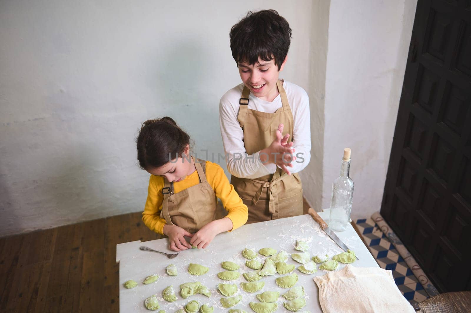 Cooking class for kids. Two kids, boy and girl preparing family dinner, standing at floured kitchen table and modeling dumplings or Ukrainian varennyky in the rural house kitchen interior. Top view by artgf