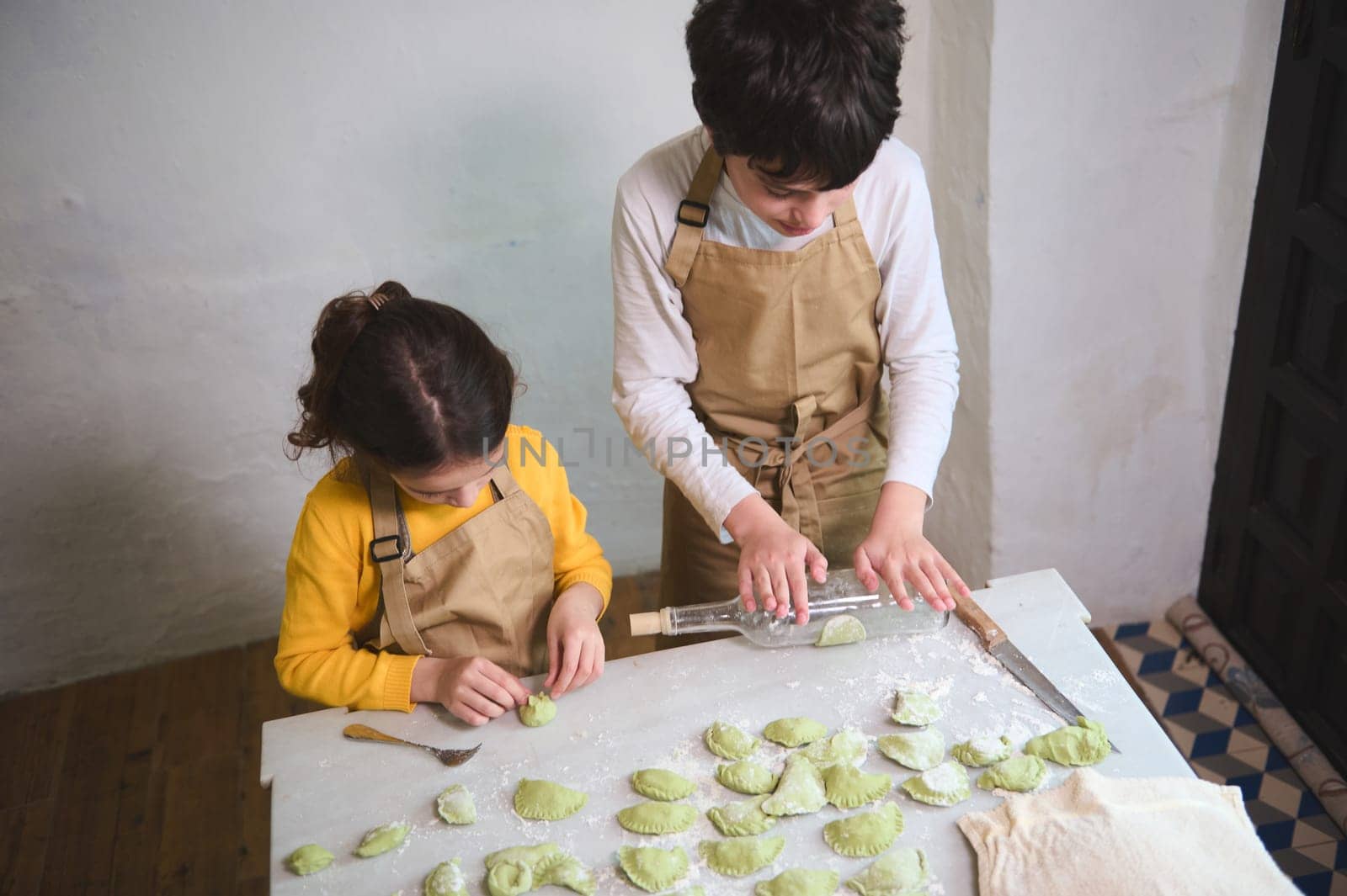 Cute children in the rural kitchen, rolling out dough with a wine bottle, sculpting dumplings with mashed potatoes filling. Cooking homemade vegetarian dumplings, Italian ravioli or Ukrainian varenyky by artgf