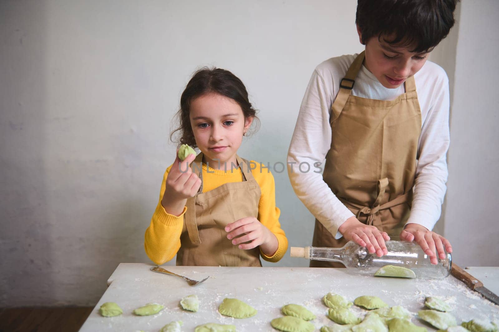 Lovely little girl stuffing and molding dough while her brother using wine bottle as rolling pin, rolling out a raw green dough. Kids making ravioli, varennyki, pelmeni against white wall background