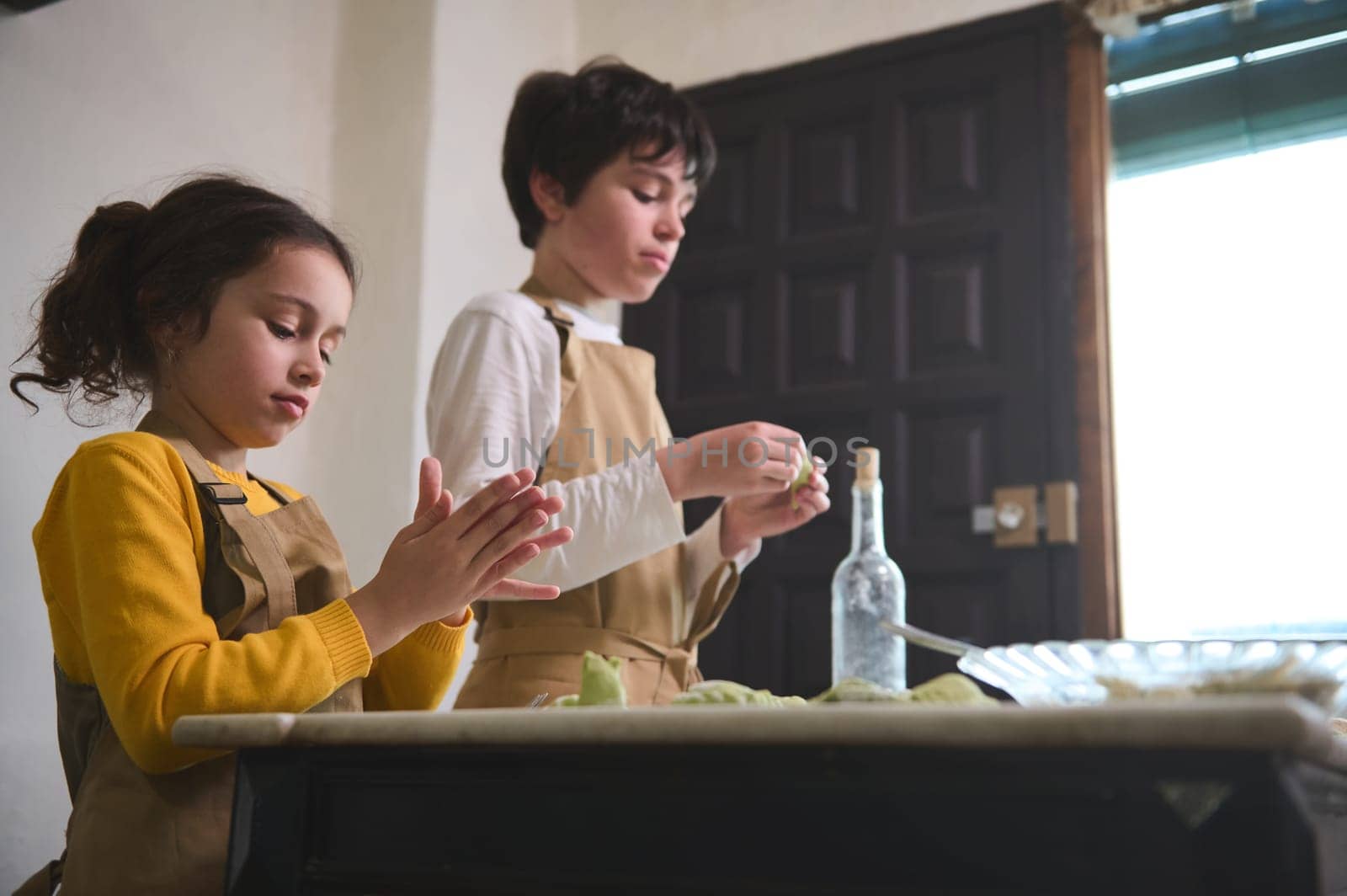 Kids modeling dumplings, standing at floured table at home kitchen interior. Caucasian boy and girl preparing a family dinner together, sculpting green ravioli according to Italian traditional recipe