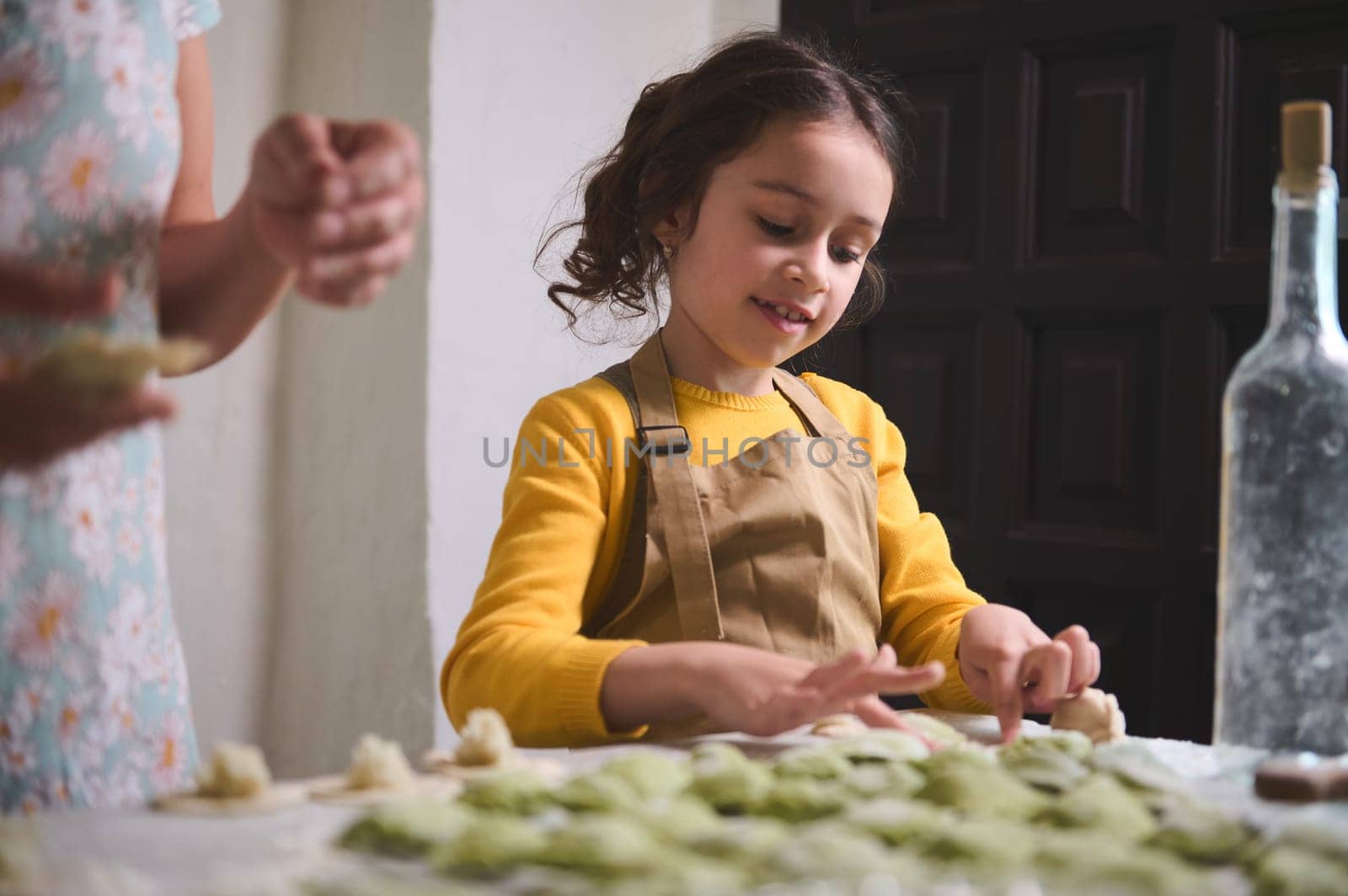 Adorable little girl in beige apron, preparing dumplings with her mother, standing at floured table in the rustic kitchen of rural house. Family traditions and culture. Cooking varennyky or ravioli.