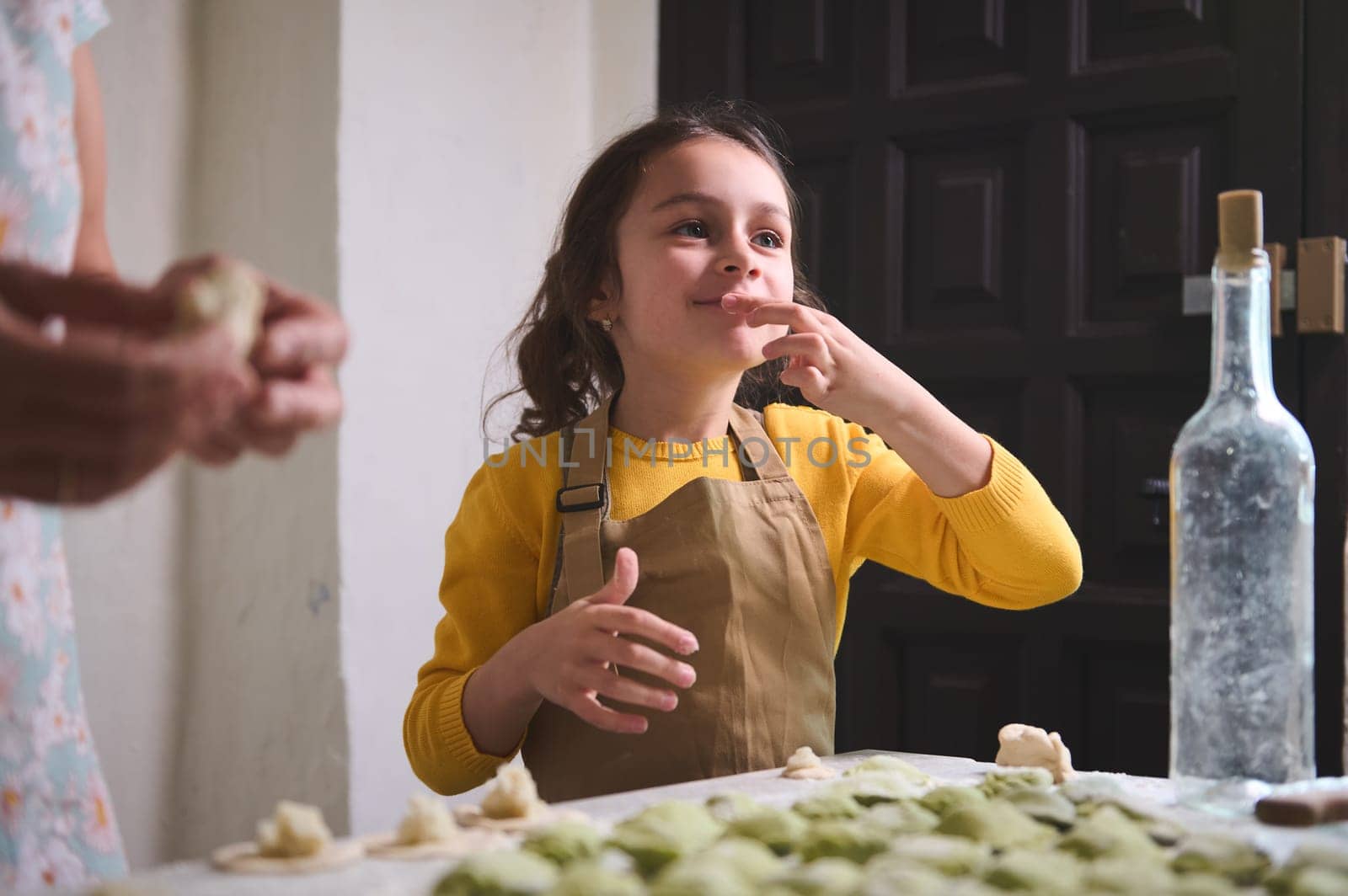 Caucasian adorable mischievous little child girl in beige apron, smiling while tasting mashed potato while cooking dumplings with her mother in the home kitchen.