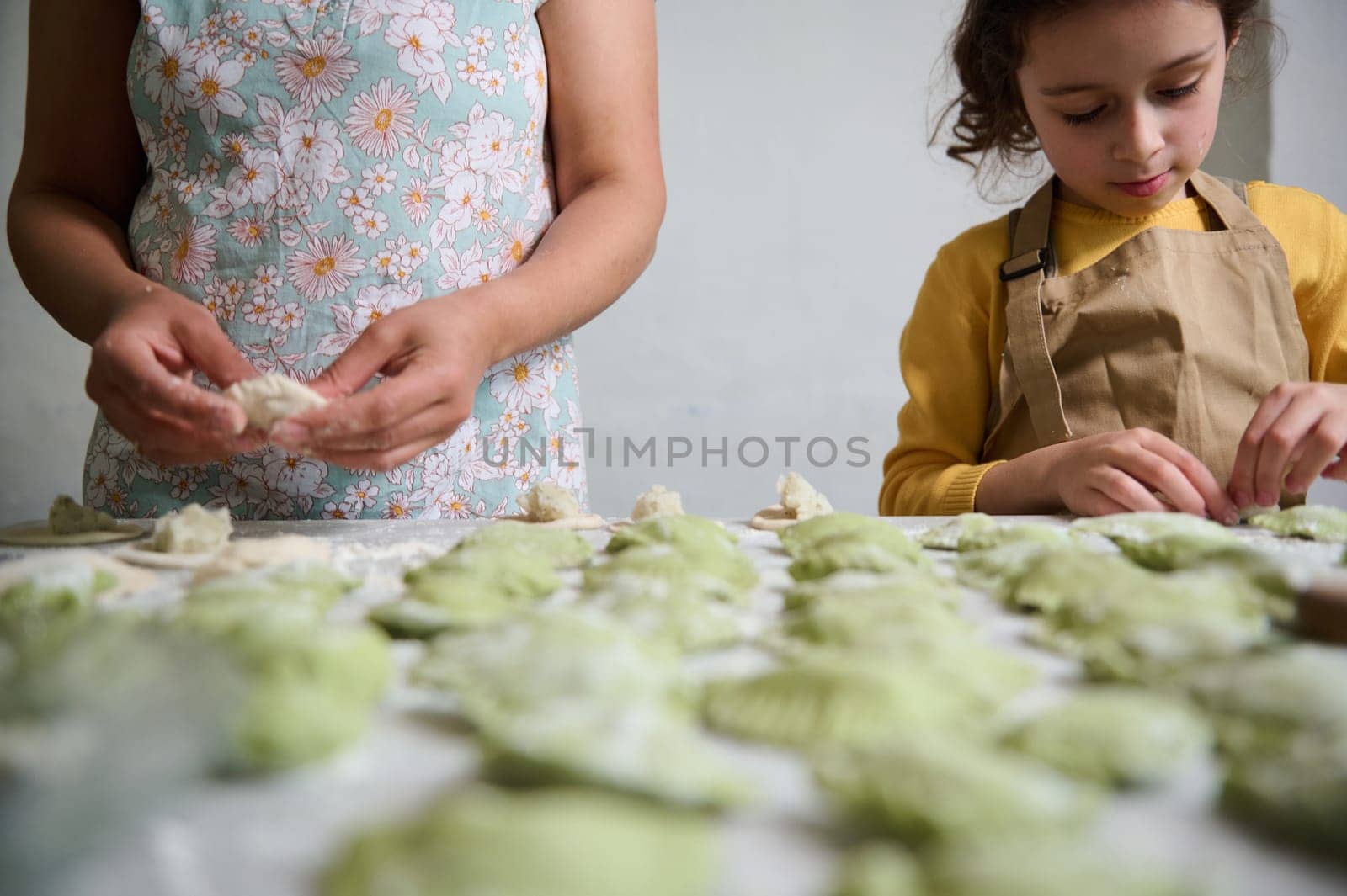 Focus on molded homemade dumplings on floured table against the background of young mom and daughter cooking together. Ravioli. Pelmeni. Varennyky stuffed with mashed potatoes by artgf
