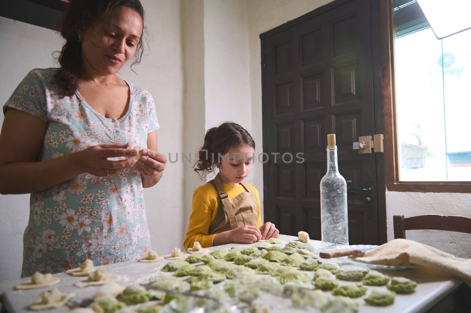 Authentic young pretty woman, mother and her little kid, daughter in the rural kitchen, sculpting dumplings from dough with mashed potatoes filling. Cooking delicious homemade vegetarian dumplings by artgf