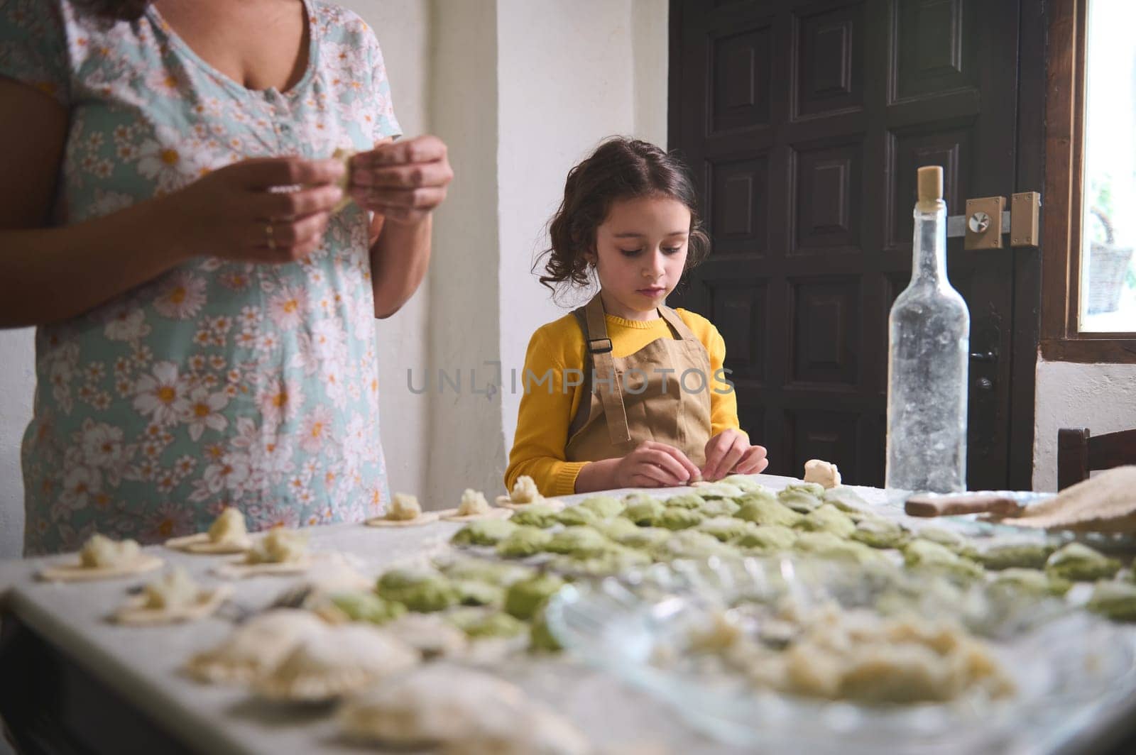 Lovely kid girl in beige chef apron, molding dumplings with mashed potato filling, standing at floured table near her mother, helping her in cuisine, learning cooking at rustic home kitchen interior by artgf