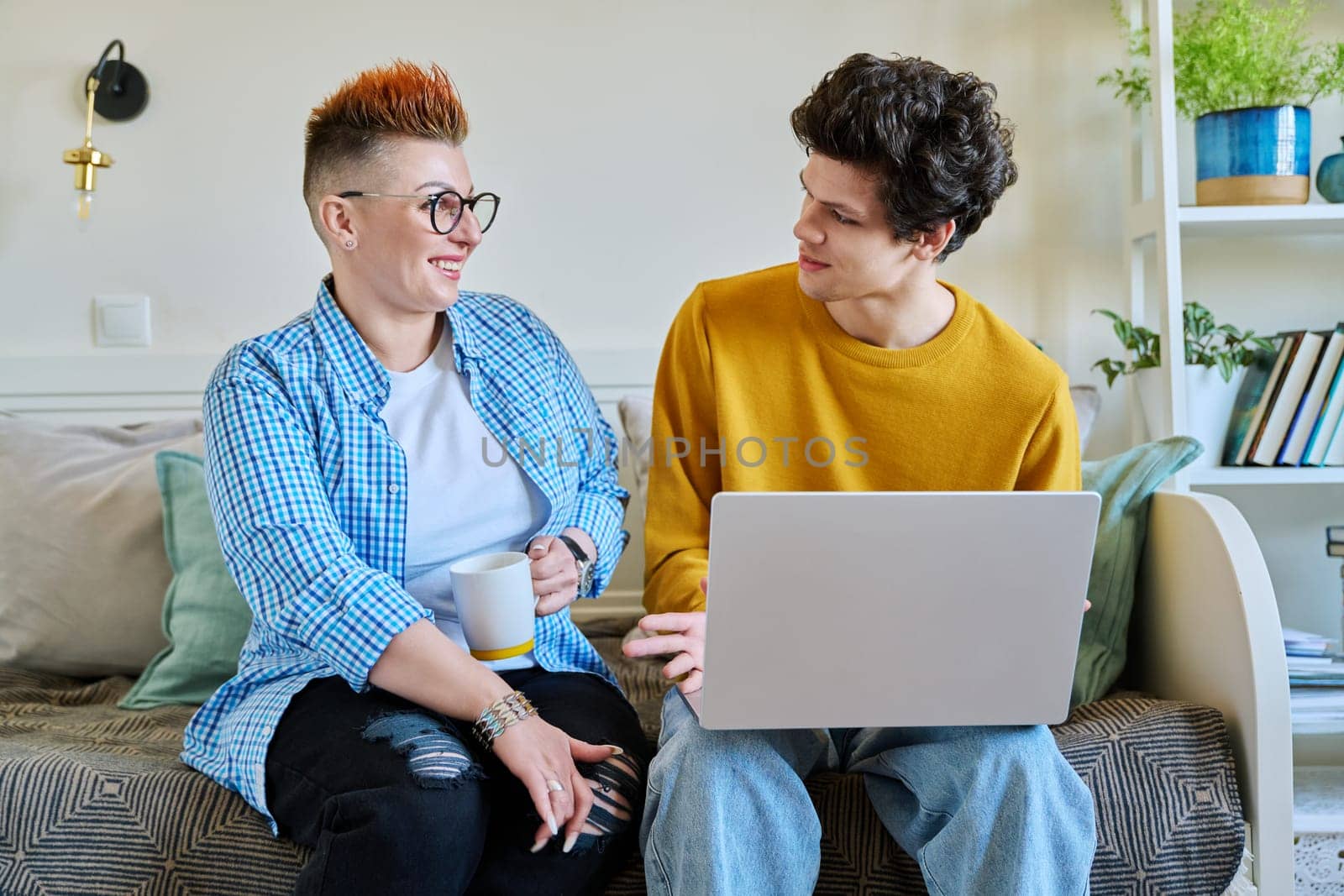 Mother and son 19-20 years old, sitting together on couch at home, looking at laptop screen by VH-studio