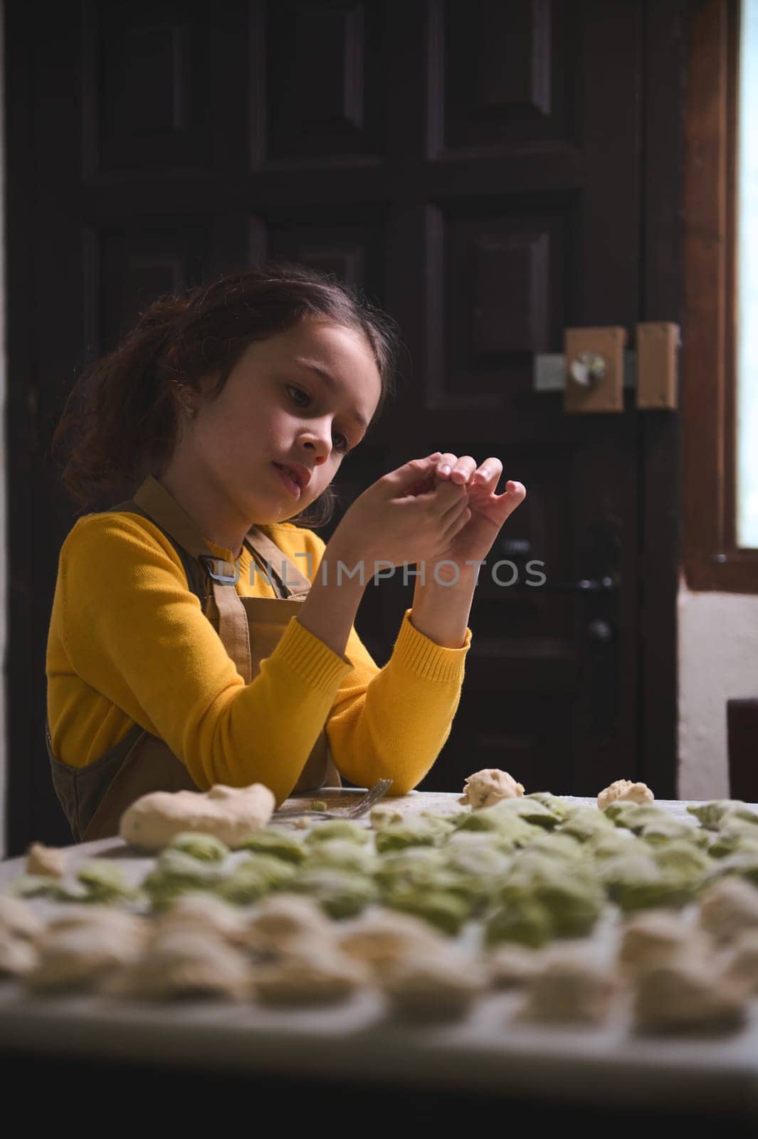 Beautiful Caucasian kid girl, little chef in beige apron, holding a homemade dumpling, standing at floured table with molded ravioli, pelmeni, vareniki, hinkali by artgf