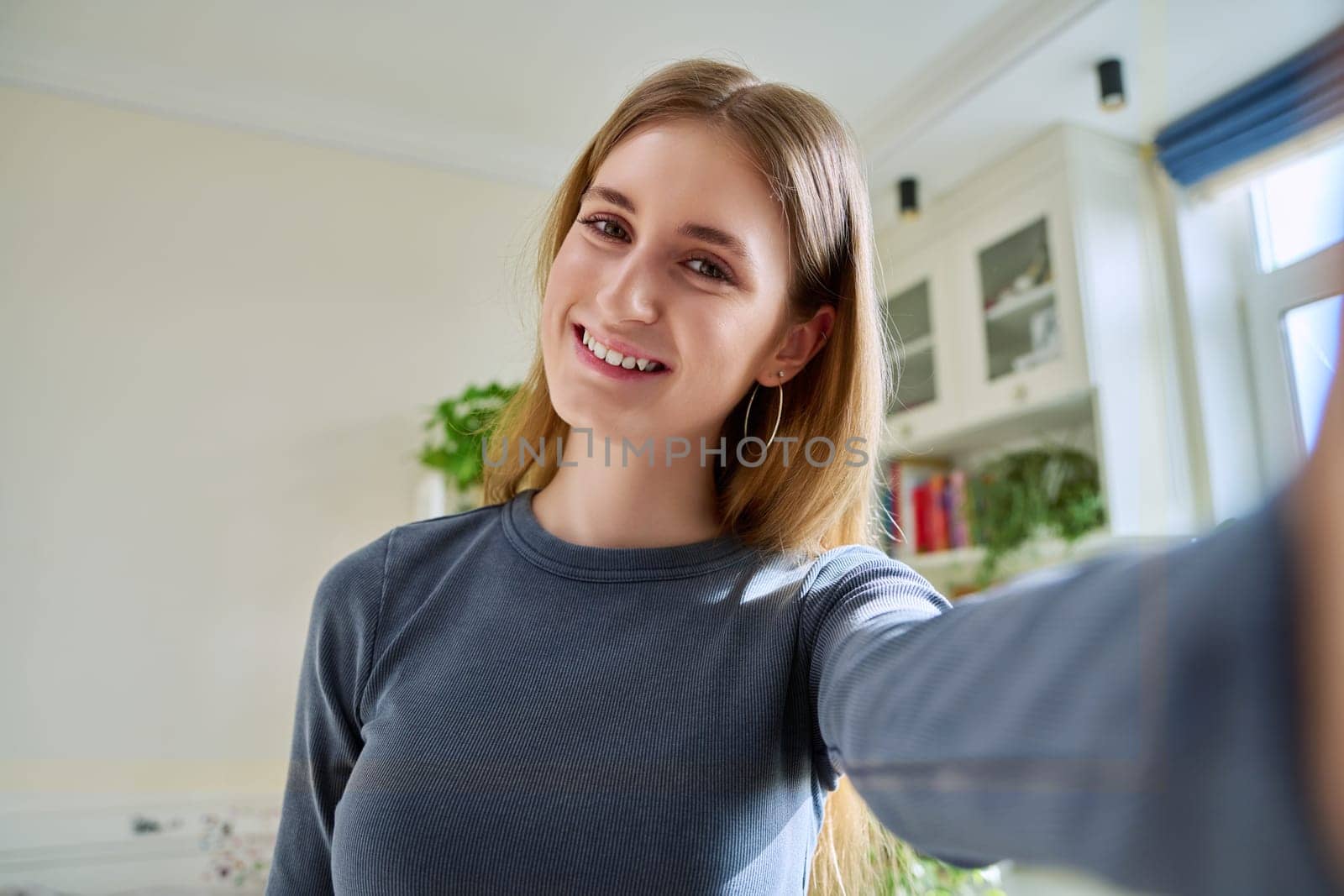 Close-up selfie portrait of teenage cheerful grimacing young female looking at web camera. Happy teenage girl 16, 17, 18 years old with blond hair, in home interior. Beauty, lifestyle, youth concept